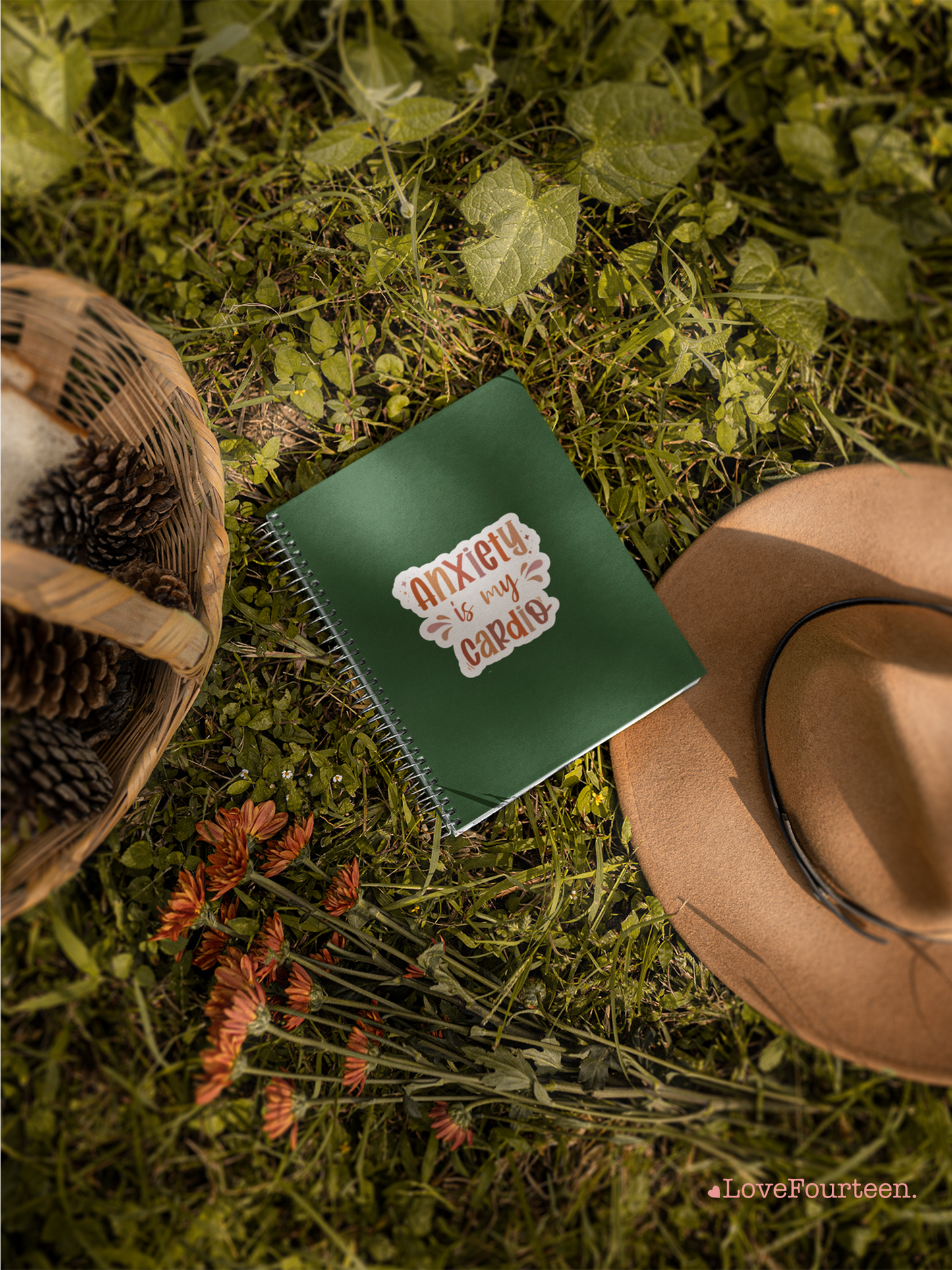 Hat, basket, and a notebook on the grass with a die cut waterproof sticker saying "anxiety is my cardio"