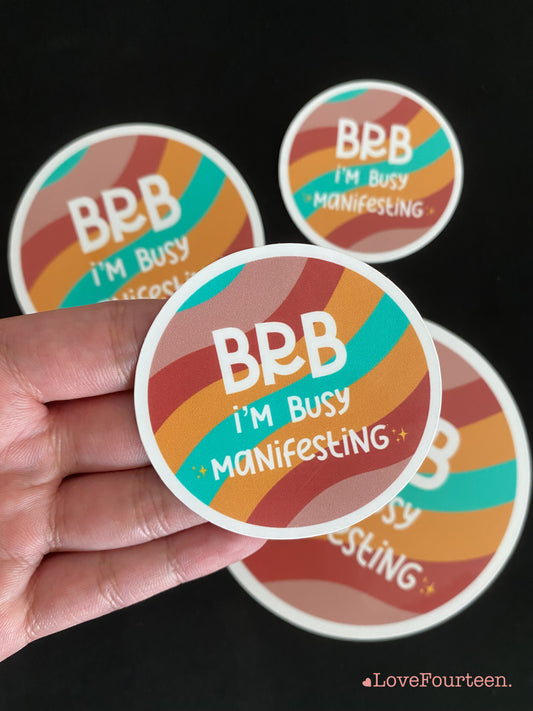 Circular waterproof die cut sticker on hand with a saying, BRB I'm busy manifesting